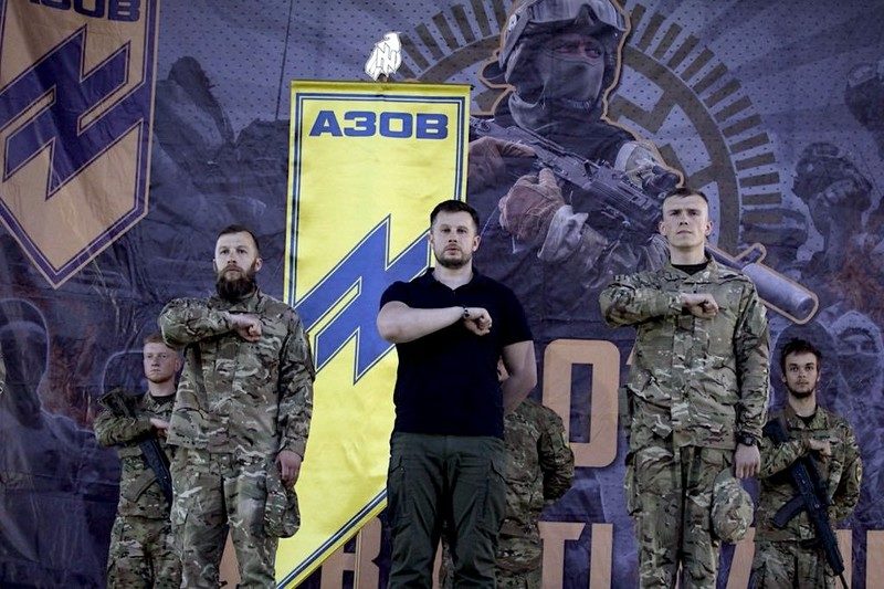 Andrij Biletskyj står foran et stort Azov-banner med uniformerte menn på begge sider. I bakgrunnen vises Azov-ss-symbolikk og militær iscenesettelse.