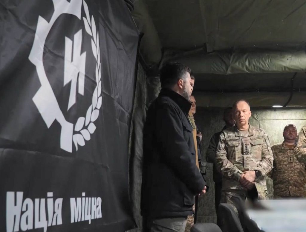 Ukrainas president Volodymyr Zelenskyj står foran en fane med et SS-lignende symbol og teksten «Нація міцна» under et besøk hos ukrainske soldater.
