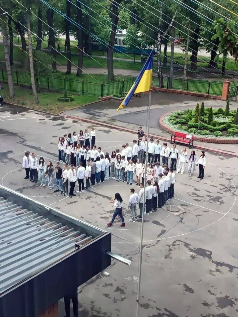 Ukrainske skoleelever kledd hovedsakelig i hvitt står samlet i en formasjon på en skolegård, ved siden av en ukrainsk flaggstang.